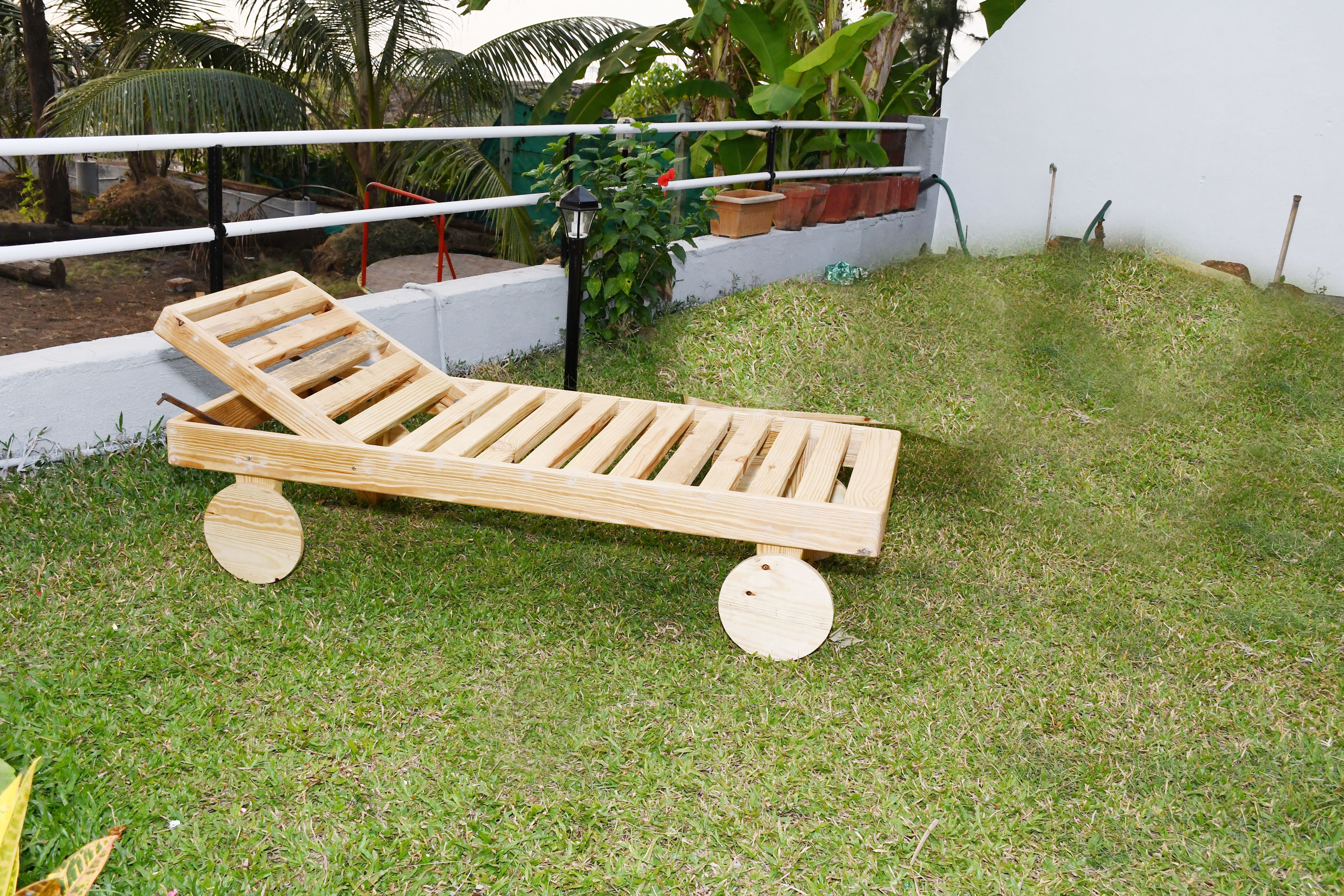 Een houten lounge stoel met een spijlenstructuur, geplaatst op een gazon met een witte muur en potplanten op de achtergrond.