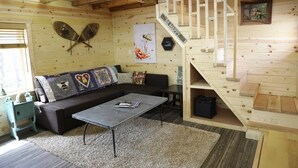 A cozy cabin interior with a wooden staircase, a sofa, a coffee table, and a wall with a 'BE HAPPY' sign.