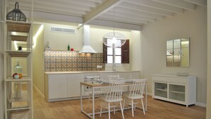 Een moderne keuken met witte kasten, een houten eettafel en stoelen. Boven de tafel hangt een lamp en rechts een witte kast met glazen deuren.
