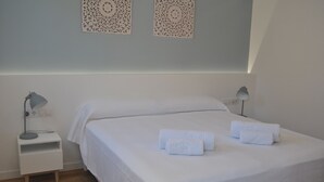 Een net opgemaakt bed met witte lakens en handdoeken, een klein nachtkastje met een lamp, en decoratieve wandkunst.