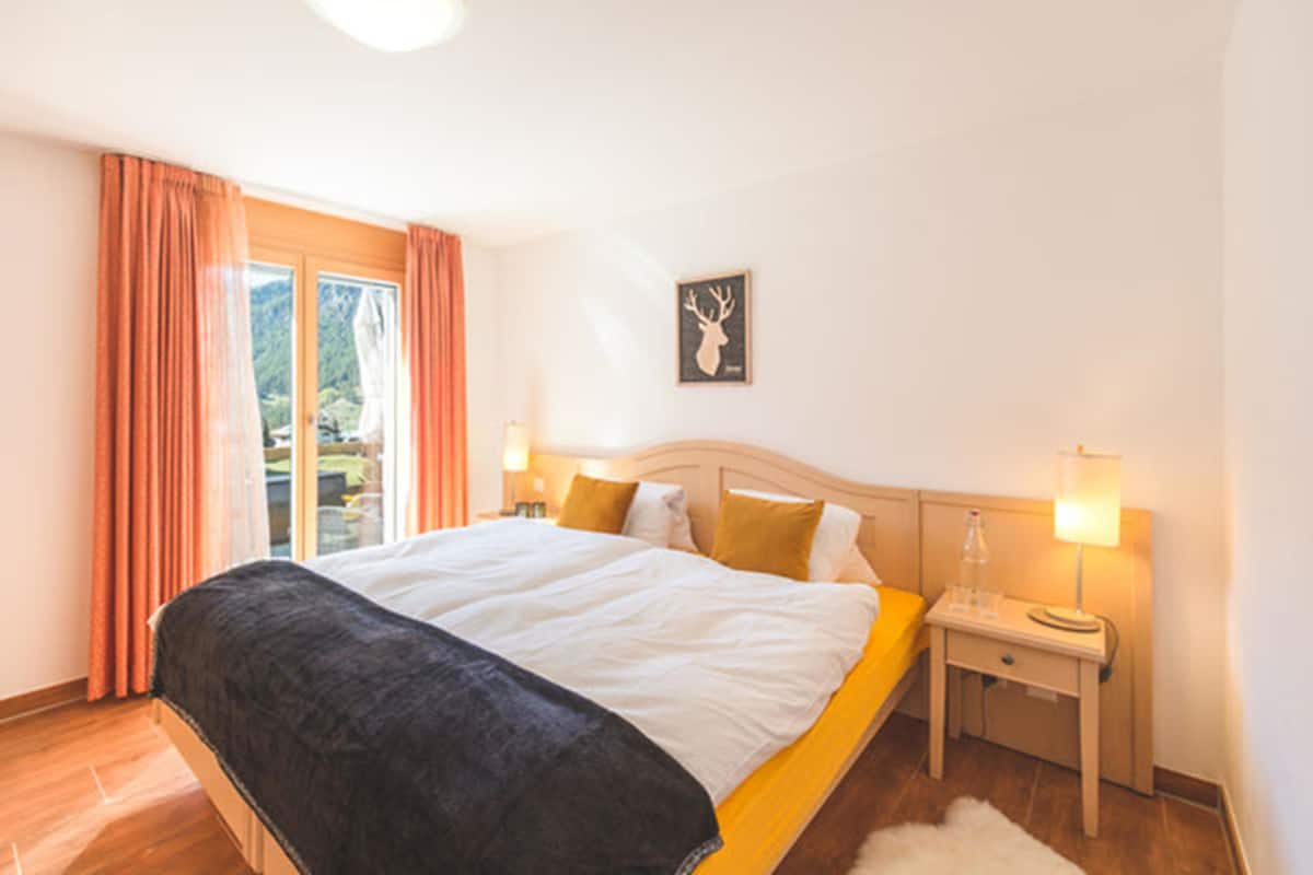 Un dormitorio con una cama, dos mesitas de noche, una mesita auxiliar, una ventana con cortinas y un cuadro colgado en la pared.
