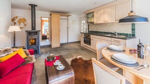 A modern living room with a red sofa, a wooden coffee table, and a fireplace. There is a kitchen area with wooden cabinets and a dining table set for two. - Chalet Alpharmonie (Zermatt)