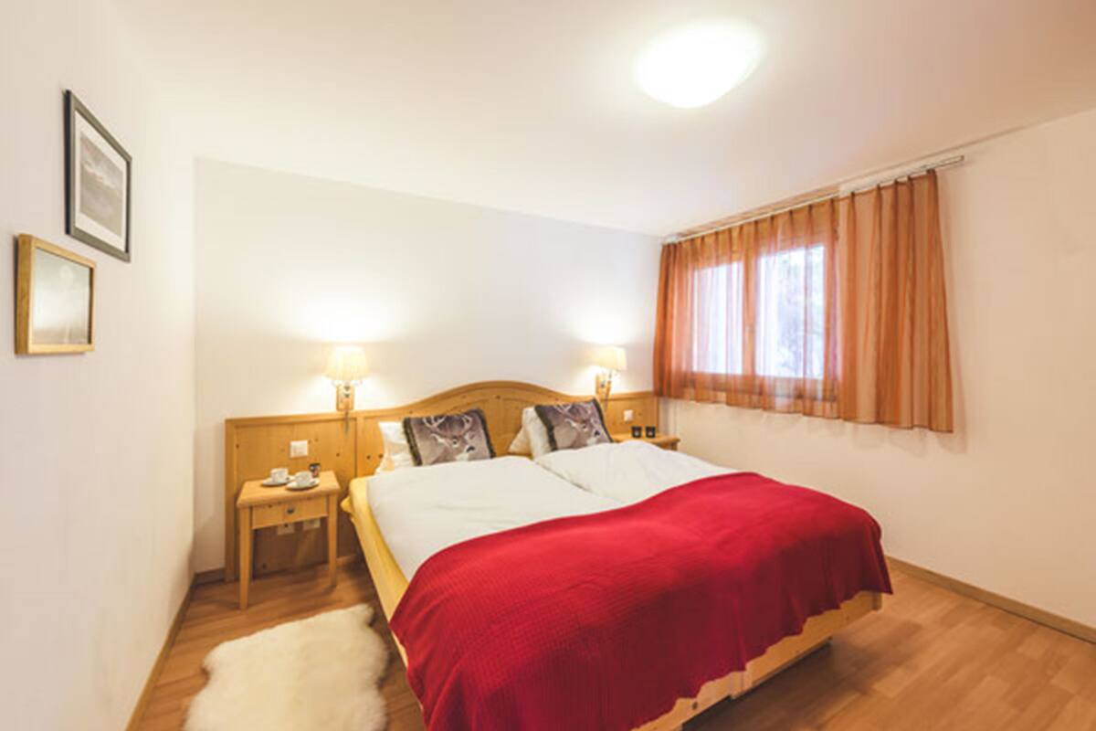 Un dormitorio con una cama de madera, dos mesitas de noche, una mesita auxiliar, una alfombra blanca y una ventana con cortinas.