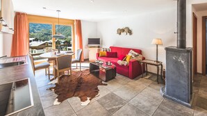 A living room with a red sofa, a stone fireplace, a dining area with a table and chairs, and a view of the mountains through large windows. - Chalet Alpharmonie (Zermatt)