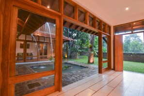 A spacious indoor area with large wooden-framed windows, a stone water feature, and a view of a lush garden. - Niketh Villa Kandy (Katugastota)