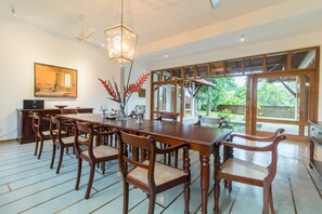 A dining area with a long wooden table and chairs, a chandelier, and large windows offering a view of greenery outside. - Niketh Villa Kandy (Katugastota)