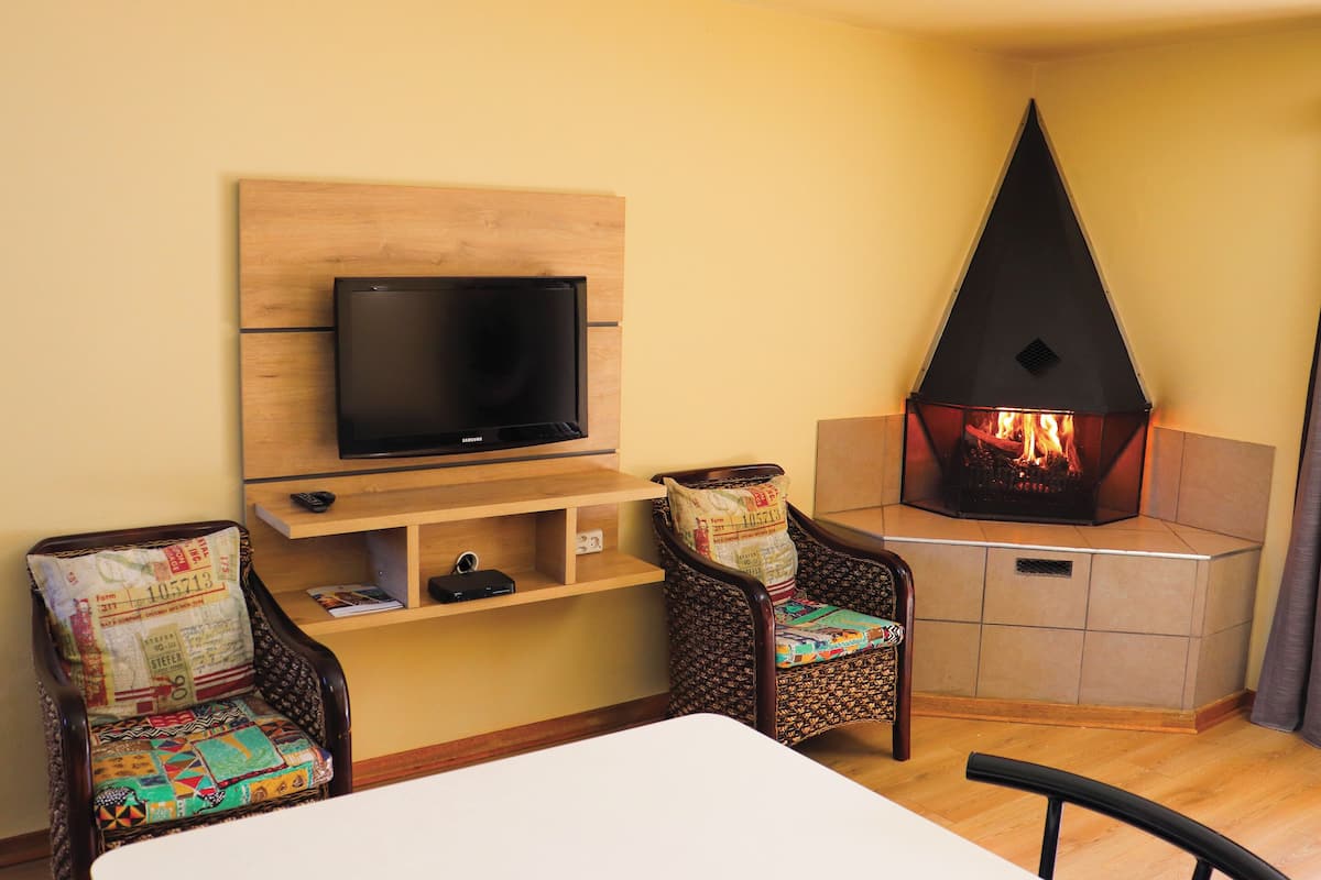 Una sala de estar con televisor de pantalla plana montado en un panel de madera, una chimenea de piedra encendida, dos sillones con estampado y una mesa.