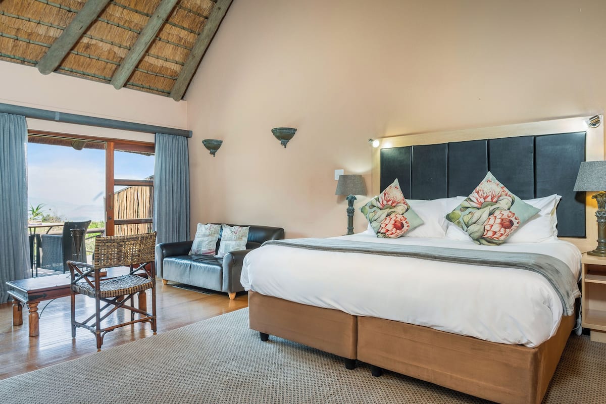 Un dormitorio espacioso con una cama grande, cabecera de madera, mesita de noche con lámpara, una silla de mimbre y vistas al exterior.