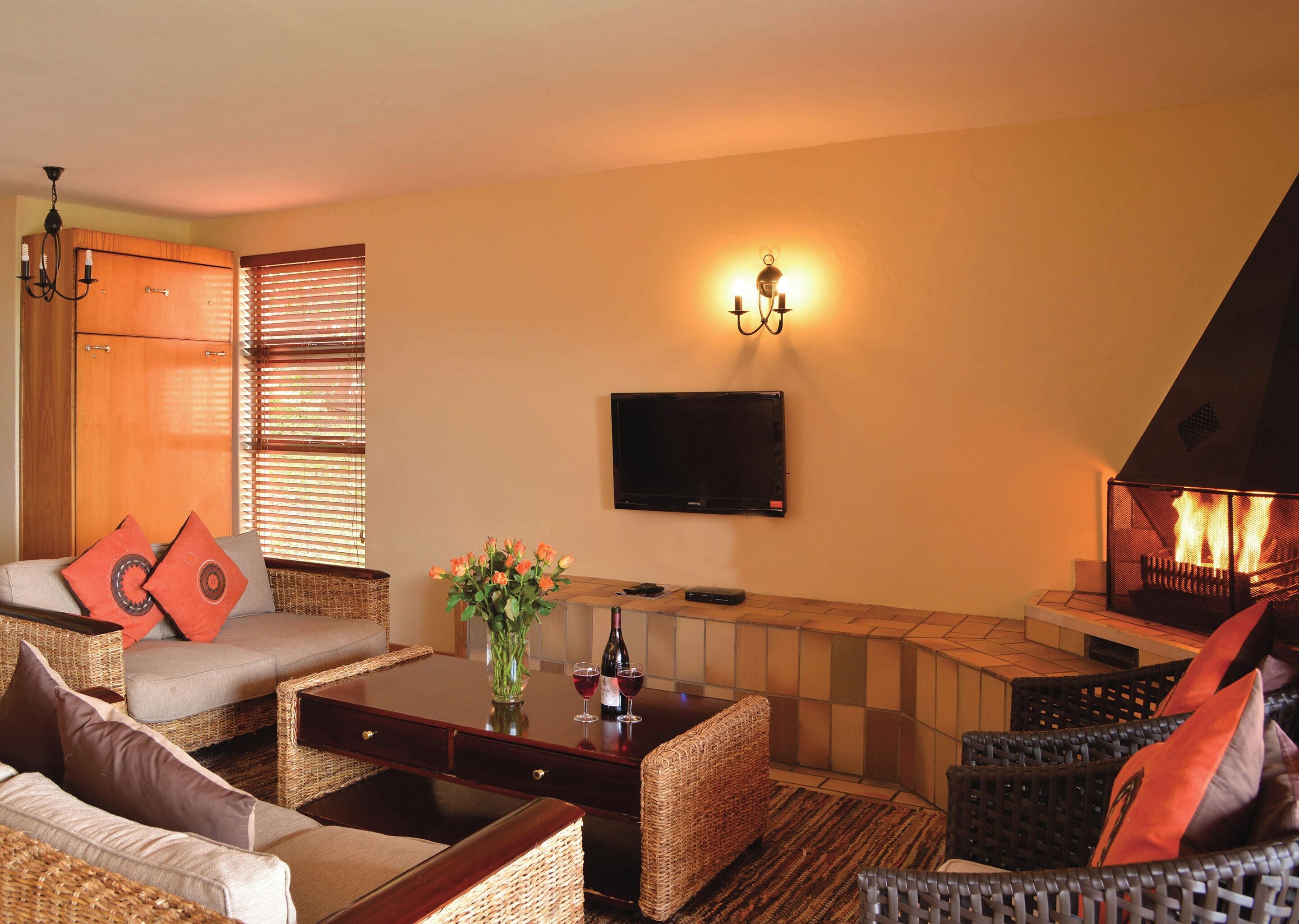 A cozy living room with wicker furniture, a flat-screen TV, a fireplace, and a wine bottle on the coffee table.