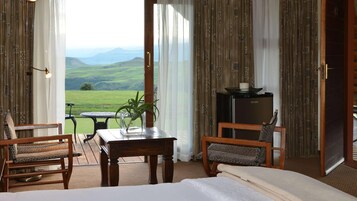 Quarto com uma cama, duas cadeiras, uma mesa com uma planta, um pequeno frigorífico e vista para colinas verdes.