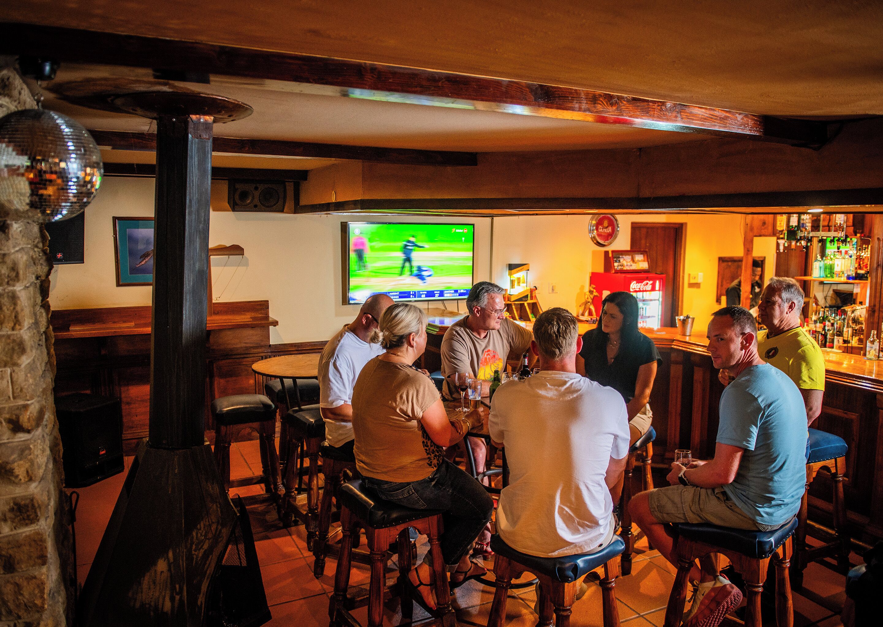 A bar area with wooden furniture, a television displaying a sports game, and a bar stocked with various bottles.