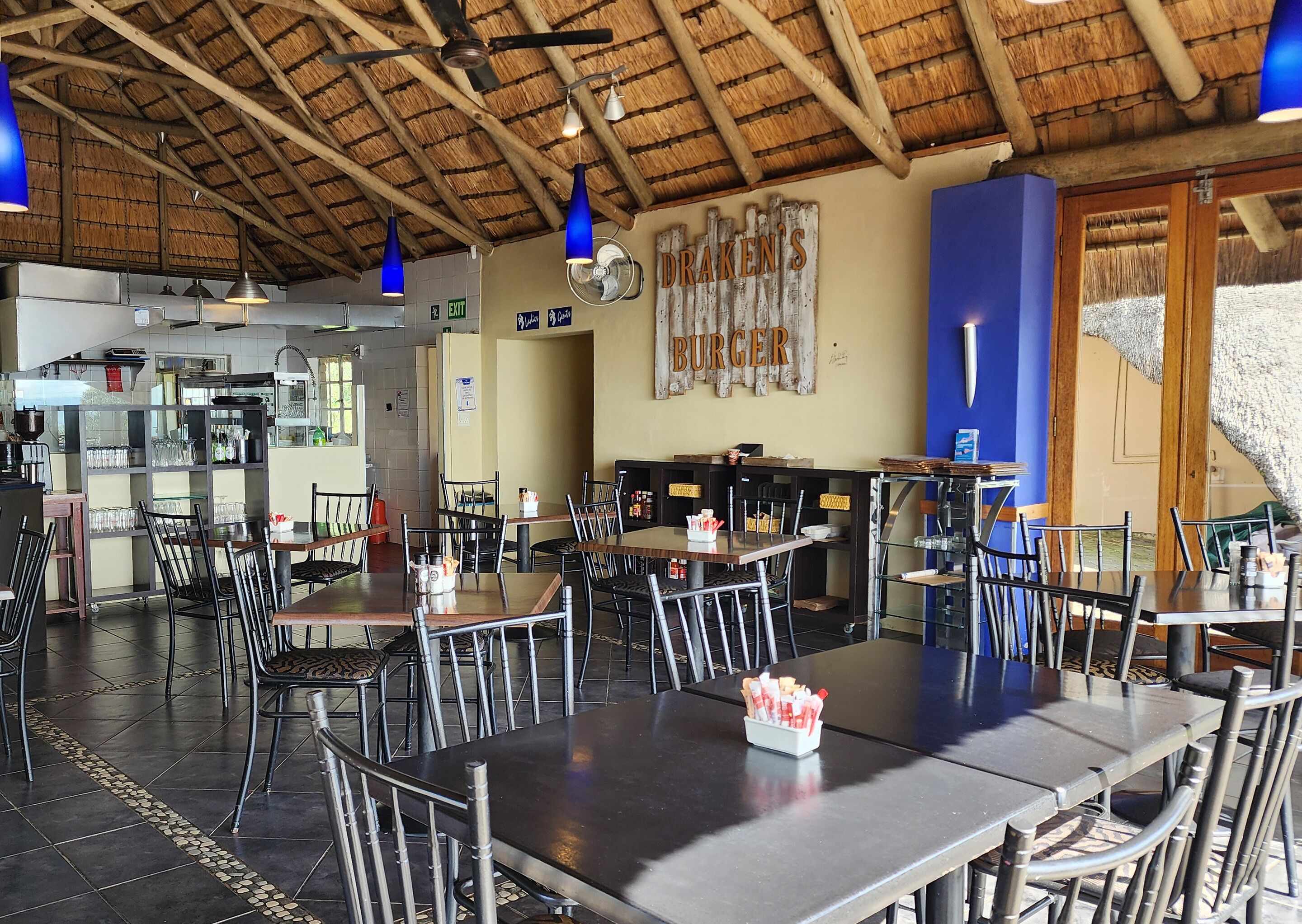 Un restaurant au décor rustique, avec des poutres en bois au plafond, des suspensions bleues et une enseigne sur laquelle on peut lire « DRAKEN’S BURGER ». Les tables et les chaises sont disposées pour accueillir les clients.