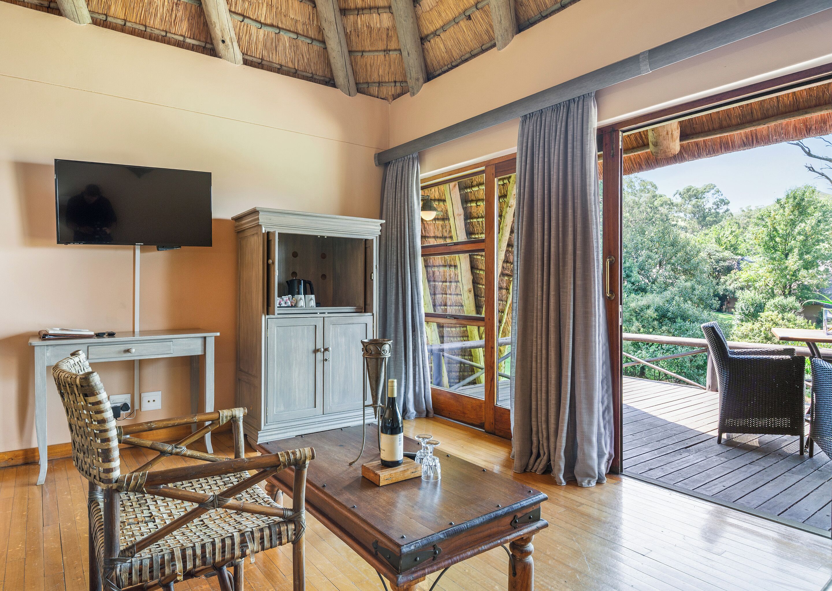 A room with a wooden desk, a television, a cabinet, wicker chairs, a table with a wine bottle, and a balcony with a view of trees.