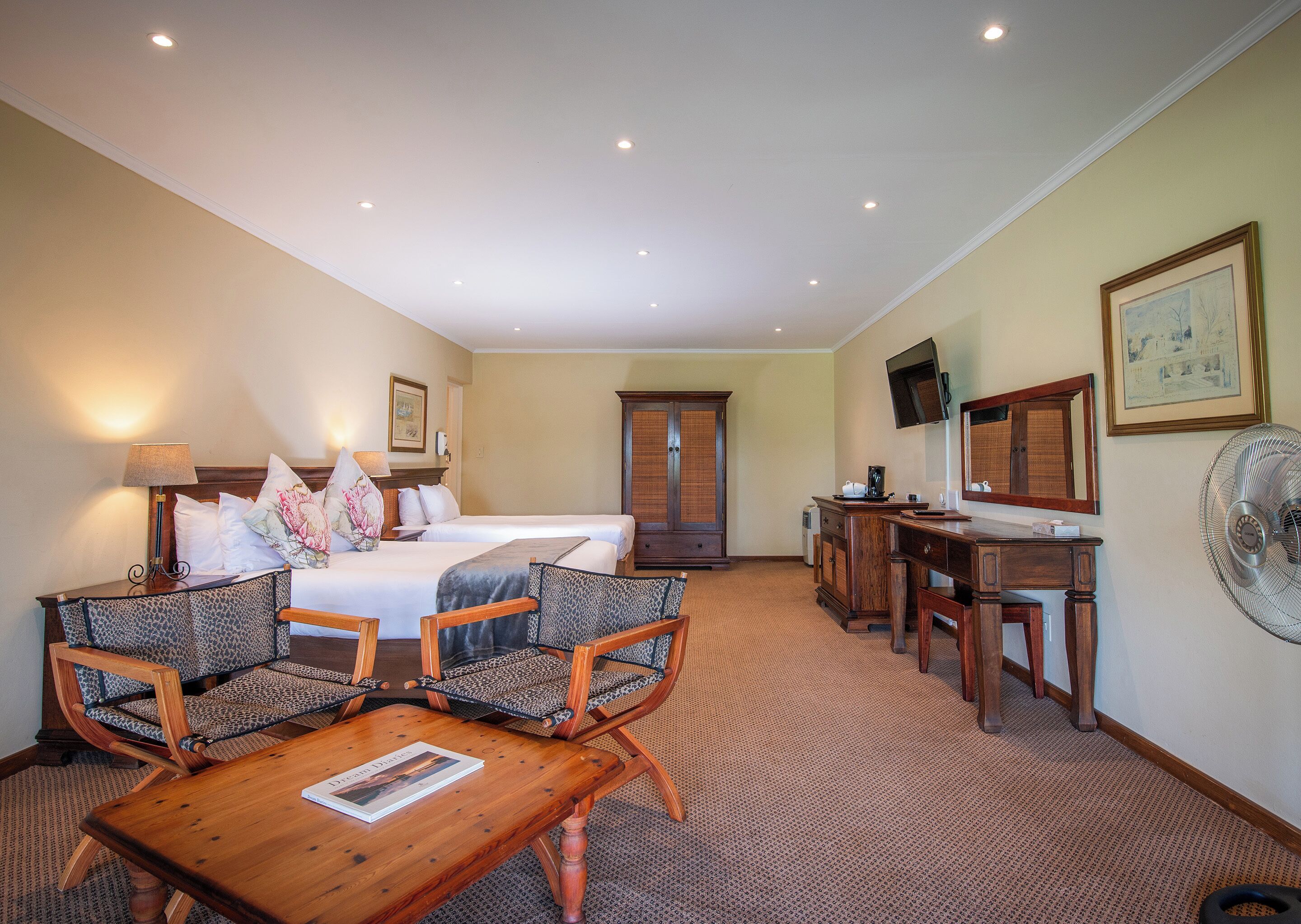 A hotel room with two beds, a desk, a chair, a table, a TV, and a fan.