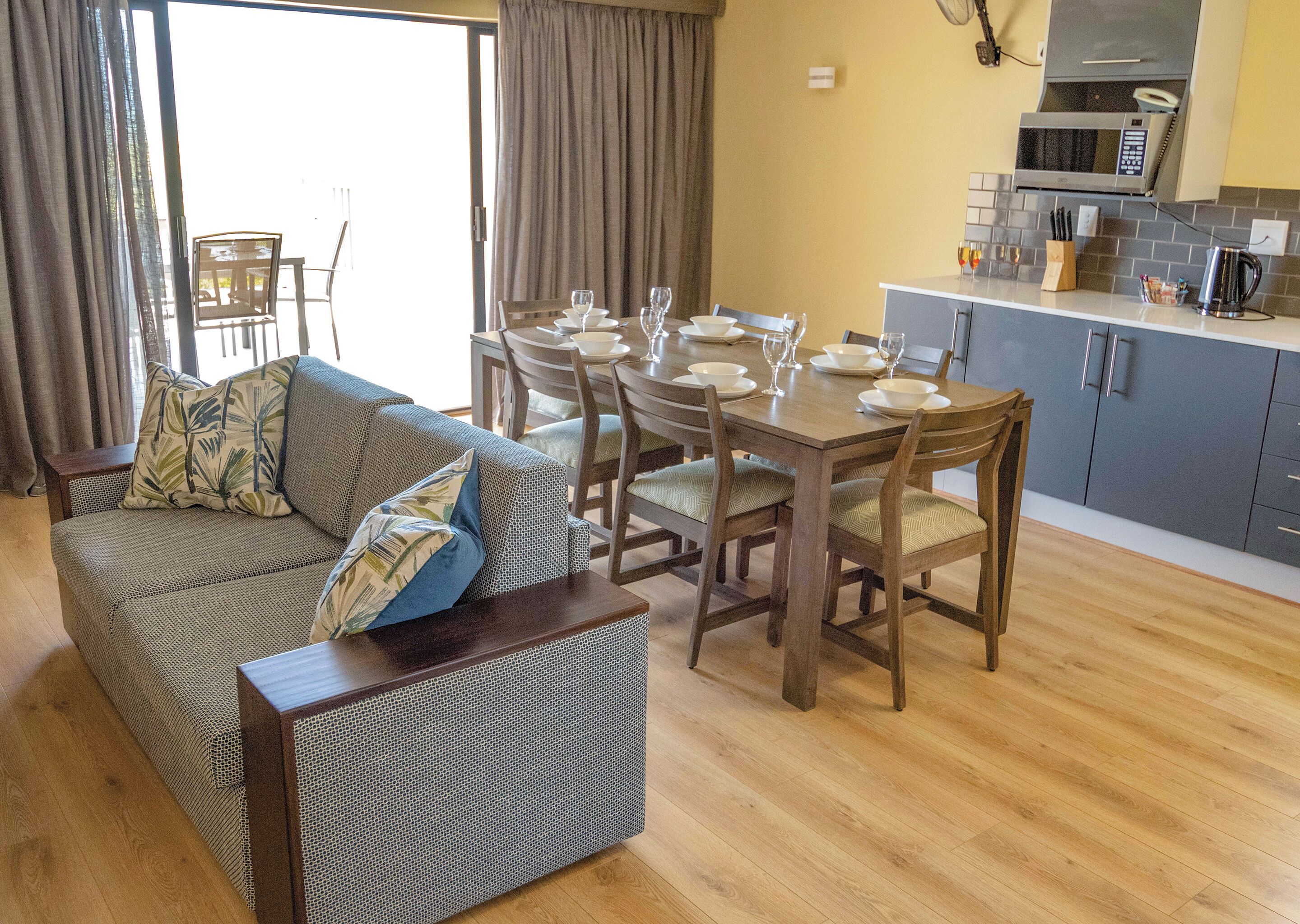 A living room with a sofa, dining table, and chairs. There is a kitchen area with cabinets and a microwave.