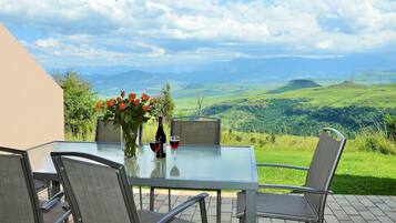 A table with chairs, a vase of flowers, and a bottle of wine, set against a scenic view of rolling hills and mountains.