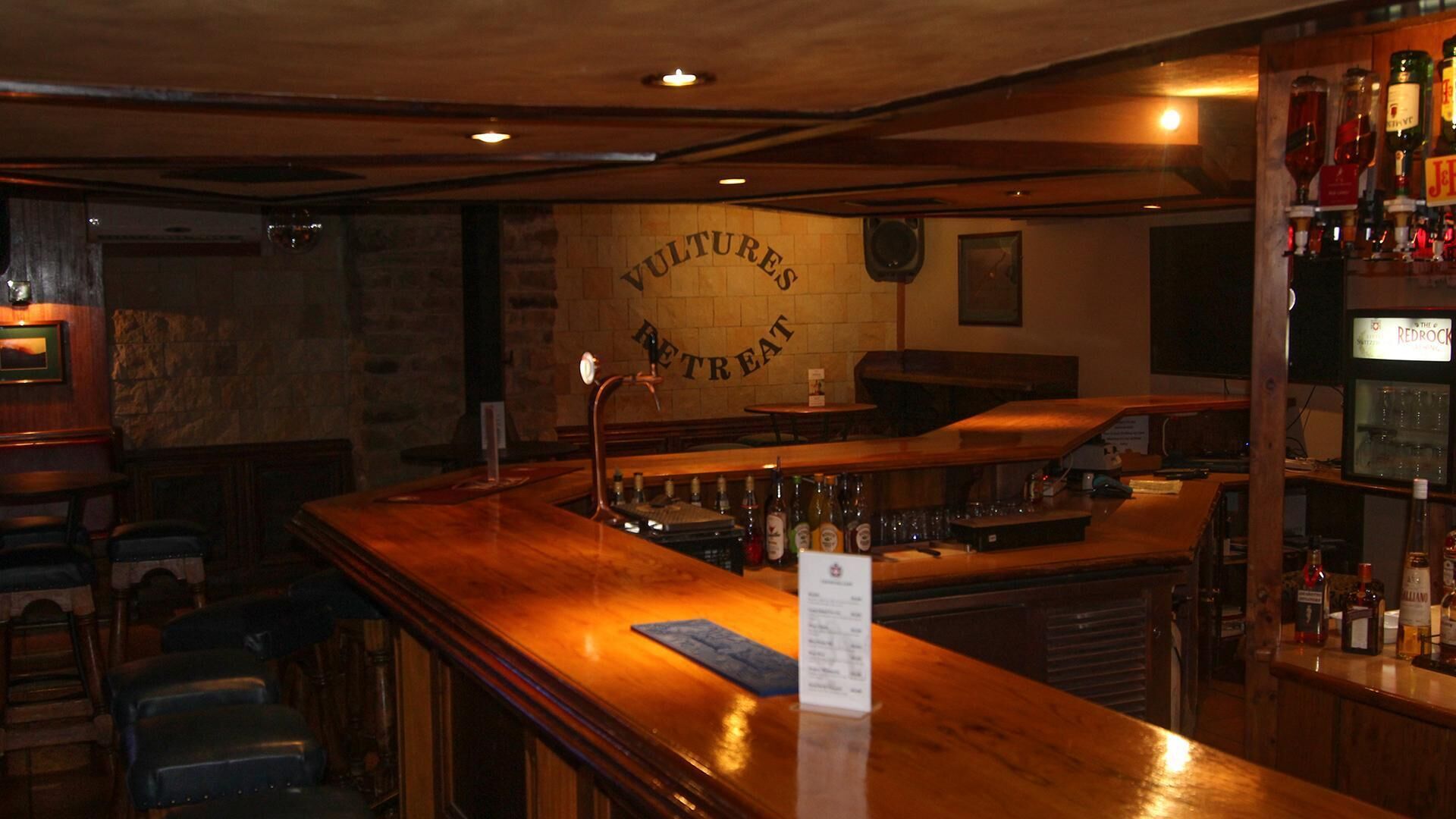 Un espace bar avec un comptoir en bois, des tabourets et une enseigne murale sur laquelle on peut lire : « VULTURES RETREAT ».