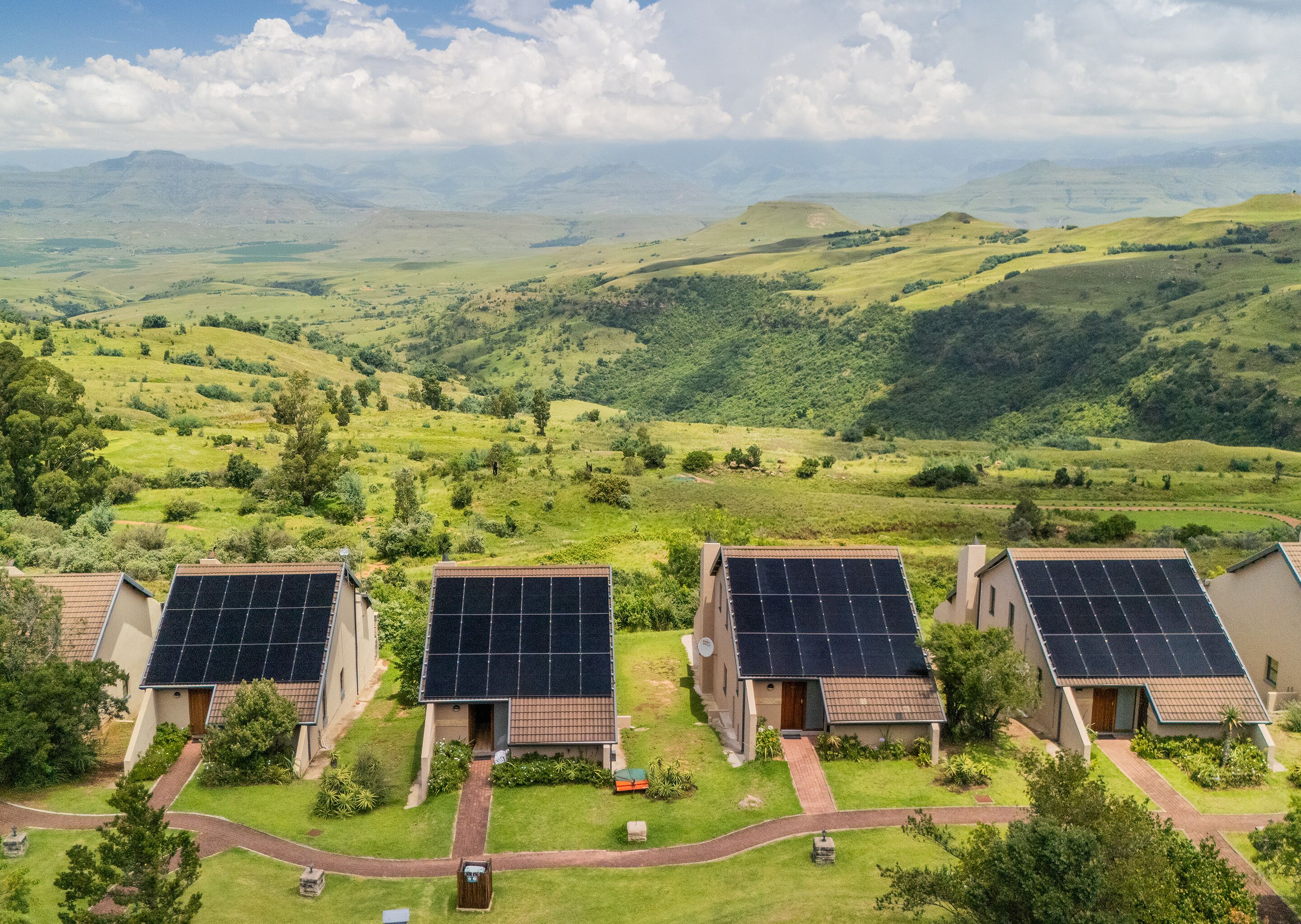 A residential area with three houses, each equipped with solar panels. The houses are surrounded by well-maintained lawns and a variety of trees.