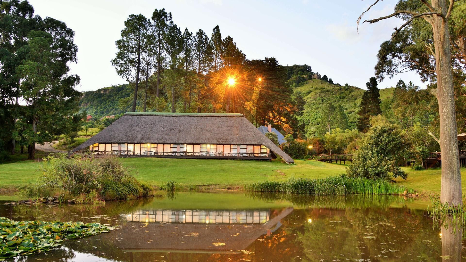 A thatched-roof building with a reflection in a pond, surrounded by trees and a hill.
