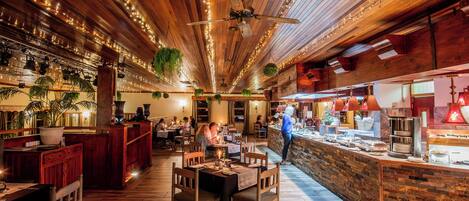 Interior de restaurante con pisos de madera, plantas colgantes, luces de cuerda y un mostrador para buffet.
