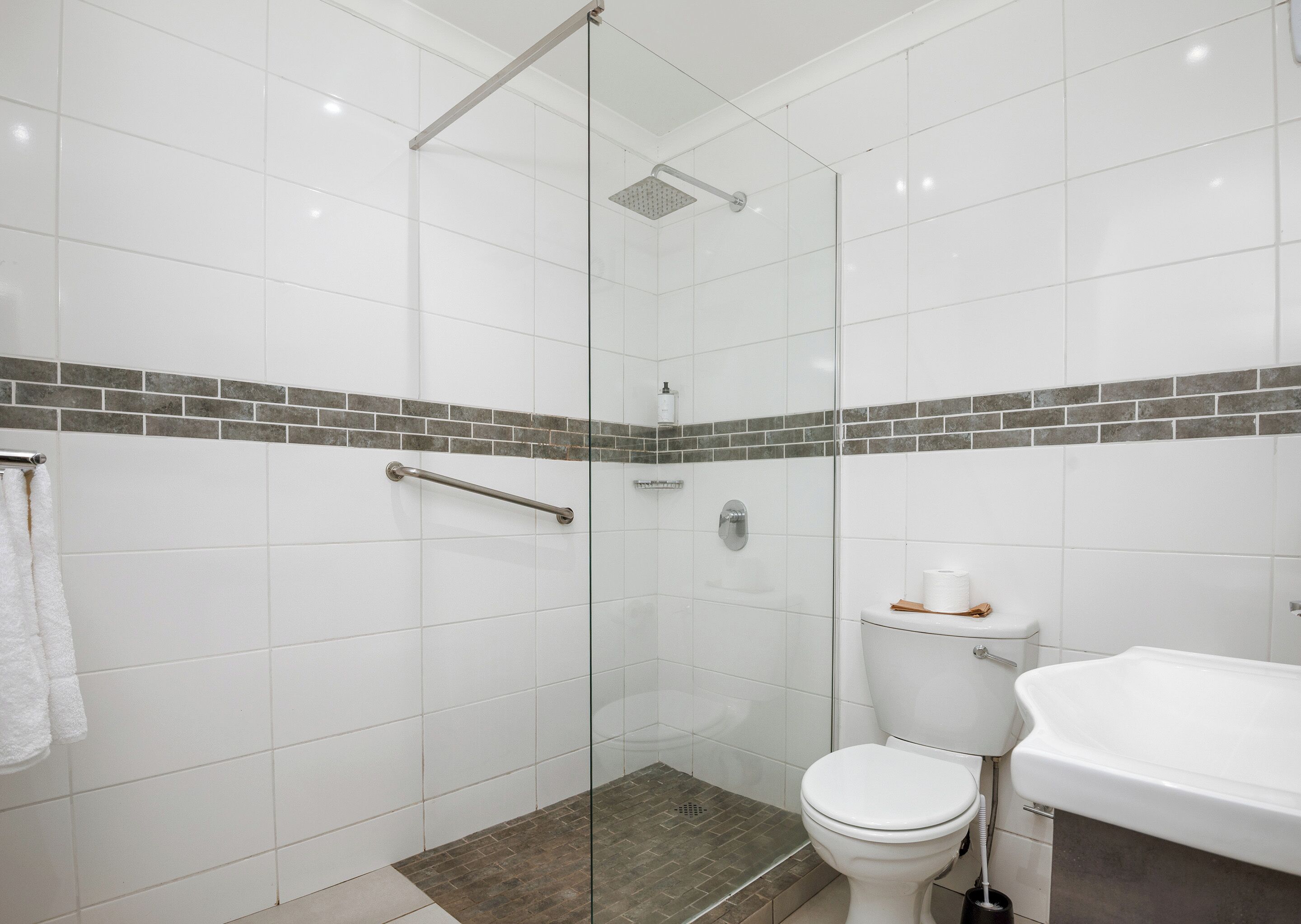 A modern bathroom with white tiles, a glass shower enclosure, a grab bar, a toilet, and a sink.