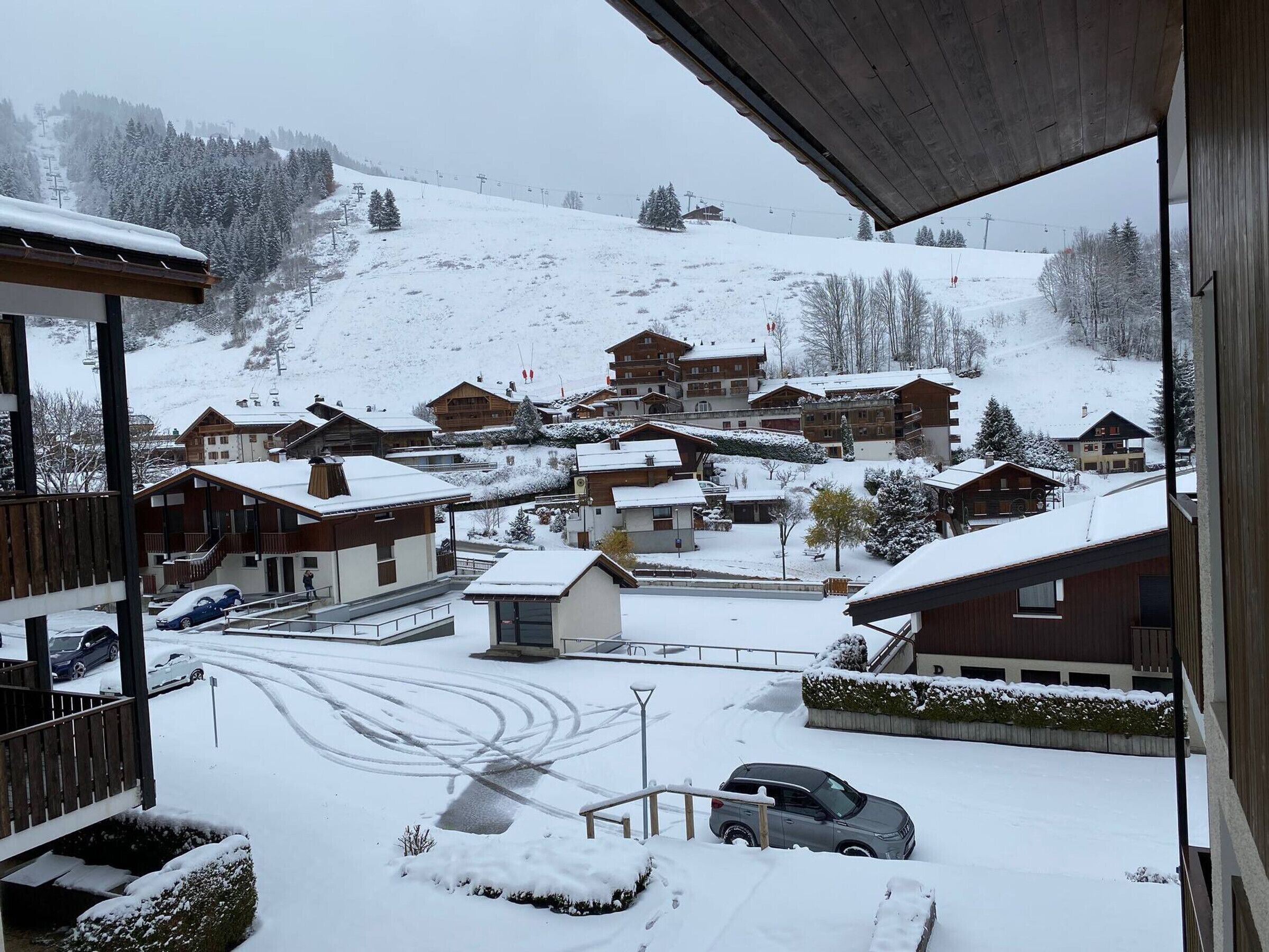 A snow-covered village with wooden buildings, a ski lift in the background, and a car parked in the foreground.