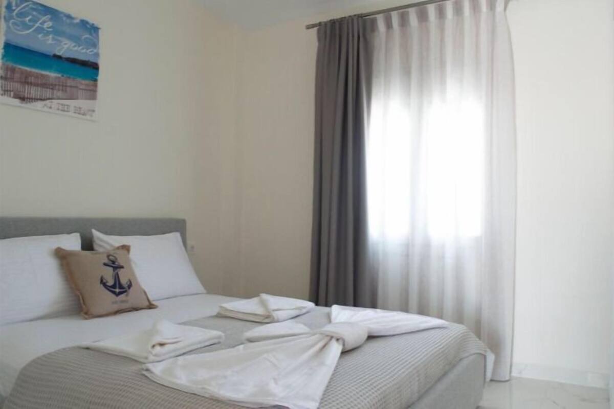 Una cama bien hecha con un cojín con diseño de ancla, una ventana con cortinas grises y una pared con un cartel.