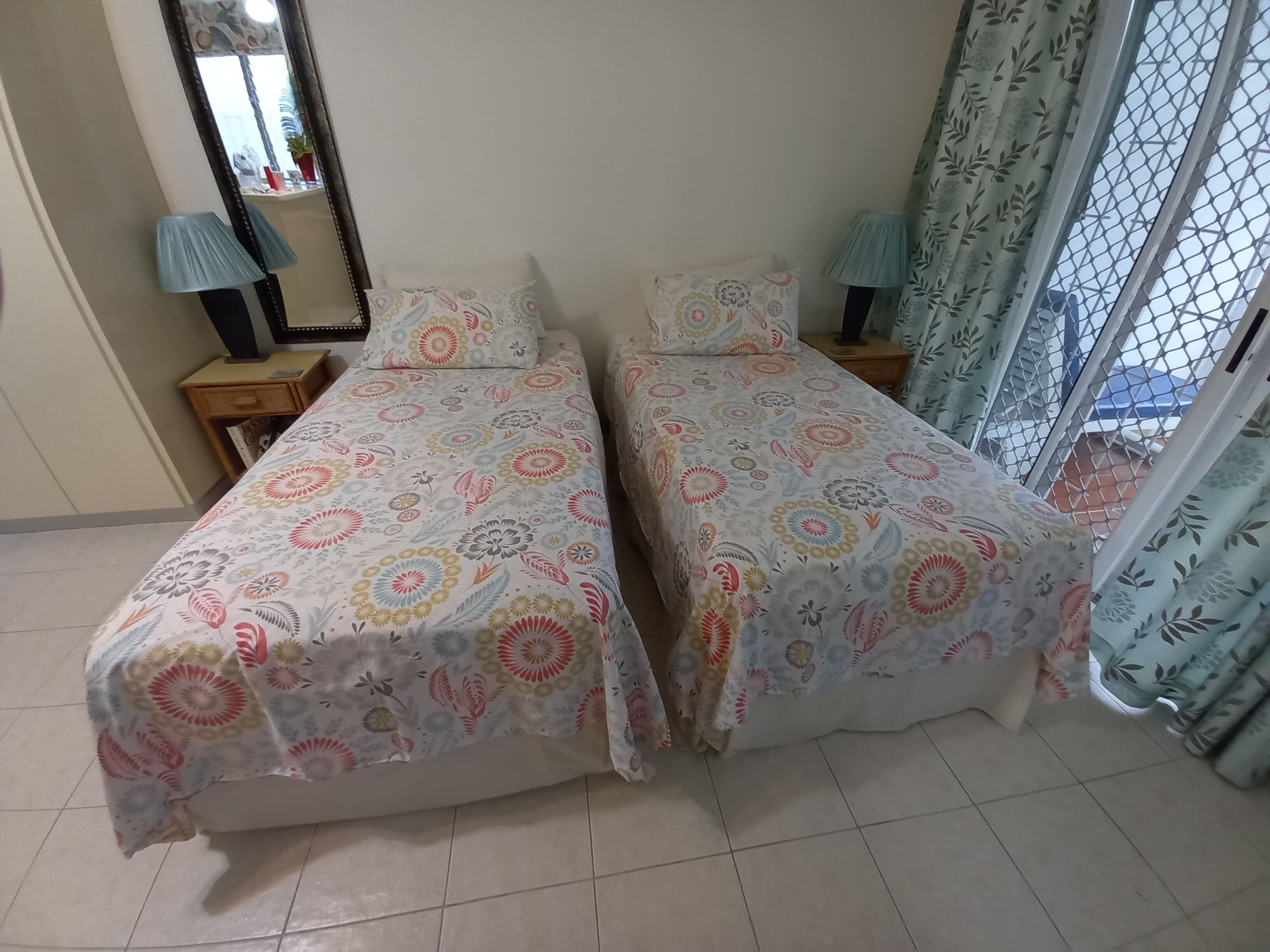 Quarto com duas camas, um espelho, uma mesa de cabeceira, um abajur e vista pela janela com cortinas.
