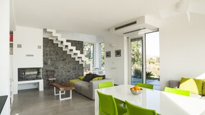 A modern living room with a stone wall, a staircase, a fireplace, and a dining area with a white table and green chairs.