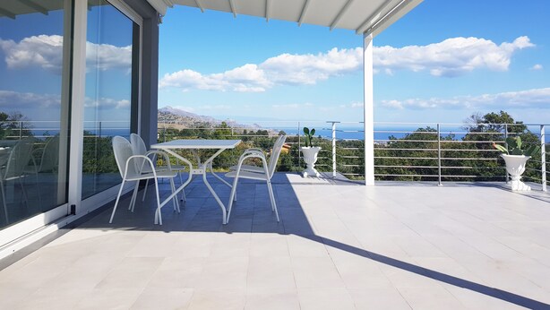 A balcony with a table and chairs, overlooking a scenic view of the sea and hills.