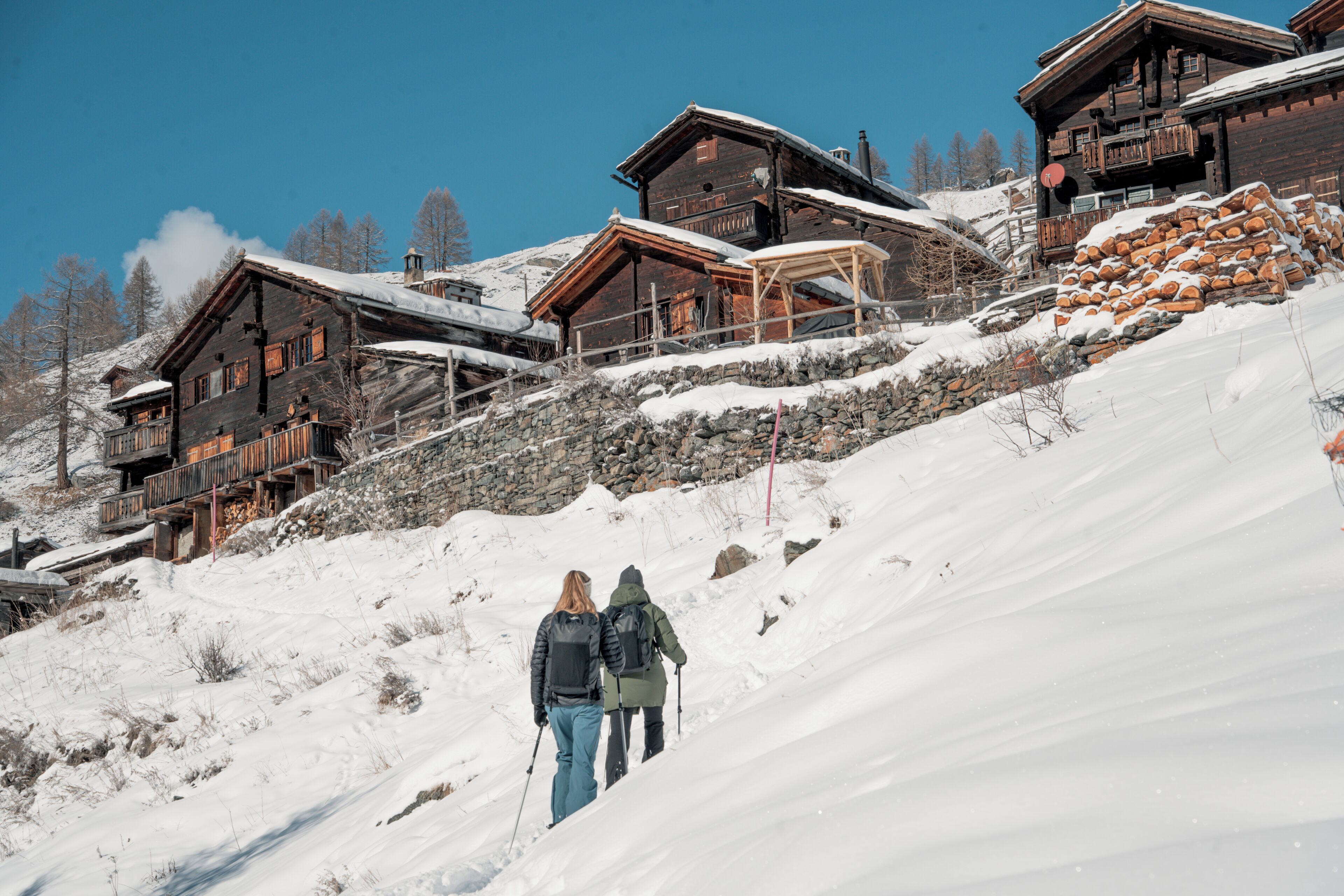 Eine Holzhütte in den Bergen mit mehreren Balkonen, einer Steinsichtmauer und einem mit Schnee bedeckten Hang.