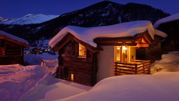 Une cabane en bois, dont le toit est recouvert de neige et dont l’intérieur est éclairé, entourée de neige et avec des montagnes en arrière-plan.