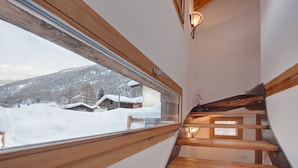 Un escalier en bois, doté d’une balustrade en verre, mène à une pièce avec une grande fenêtre donnant sur un paysage enneigé.