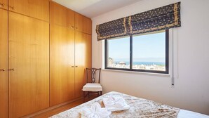 Une chambre dotée d’une grande armoire, d’un lit recouvert d’une couvre-lit à motifs et offrant une vue sur la mer par une fenêtre agrémentée d’une lambrequin à motifs.