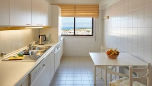 Une cuisine compacte avec des meubles blancs, une fenêtre donnant sur un beau paysage et une table sur laquelle se trouve un panier de fruits.