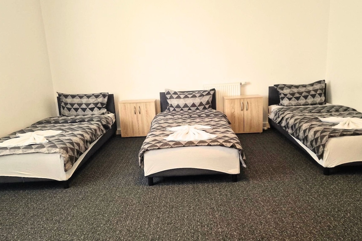 Una habitación con tres camas individuales en línea, cada una con una colcha y almohadas estampadas. Hay una mesita de noche de madera entre las camas, y una pequeña mesa con una lámpara a la derecha. El suelo está alfombrado y las paredes son lisas.