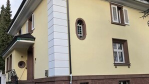 A two-story building with a yellow facade, brown trim, and a brick base. There are two windows with shutters, a small circular window, and a door with a circular window above it. - Villa Birkenfeld (Birkenfeld)
