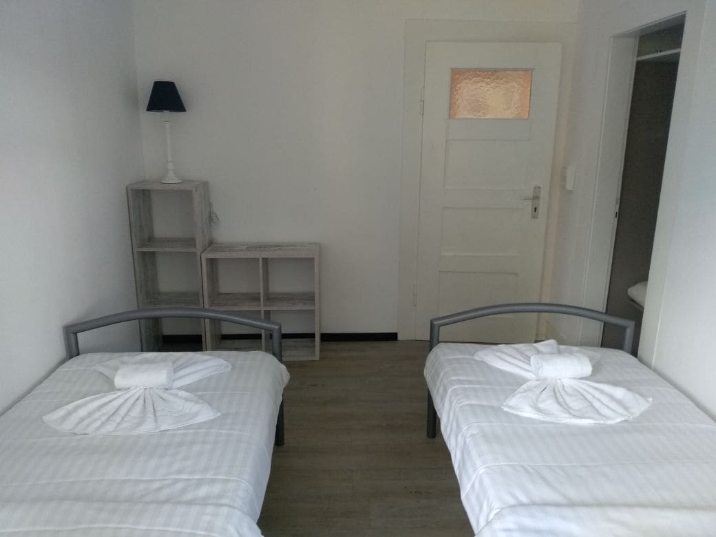 A small, clean room with two single beds, a small table with a lamp, and a door with a window.