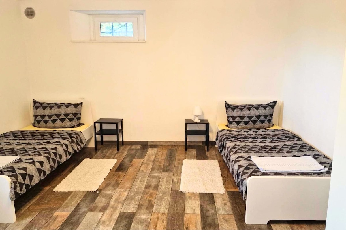 Una habitación con dos camas, cada una con ropa de cama estampada, una mesita de noche pequeña con una lámpara, un ventanal sobre las camas y suelo de madera.