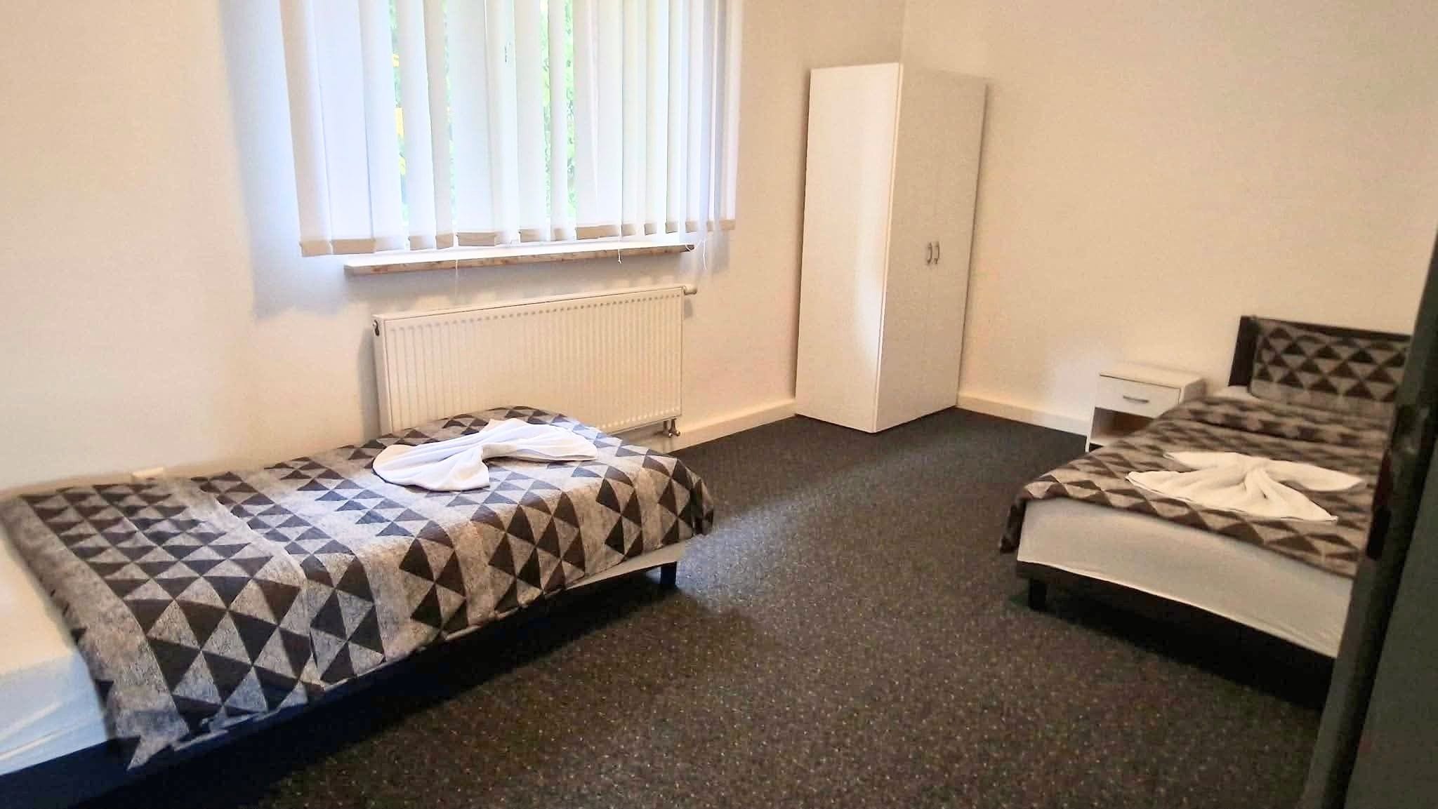 A small, sparsely furnished room with two beds, a radiator, a window with blinds, and a door.