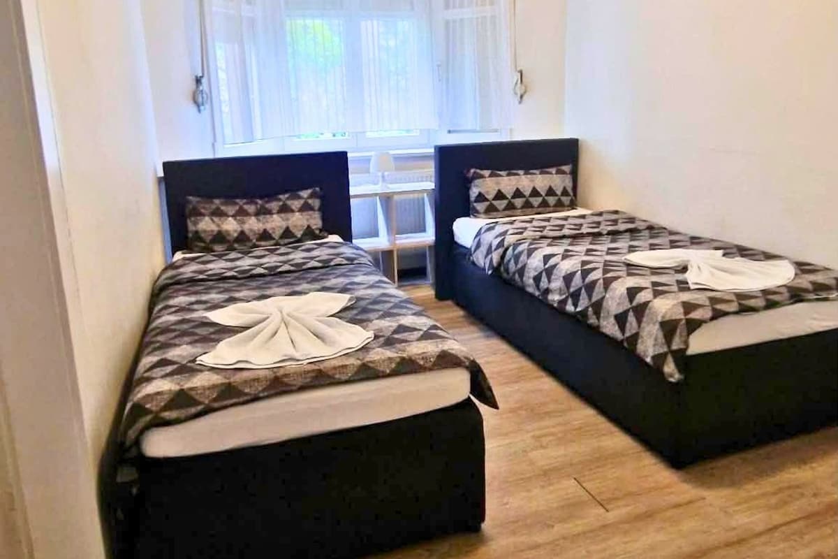 Una habitación con dos camas, un ventanal con cortinas traslúcidas y una lámpara en el techo.