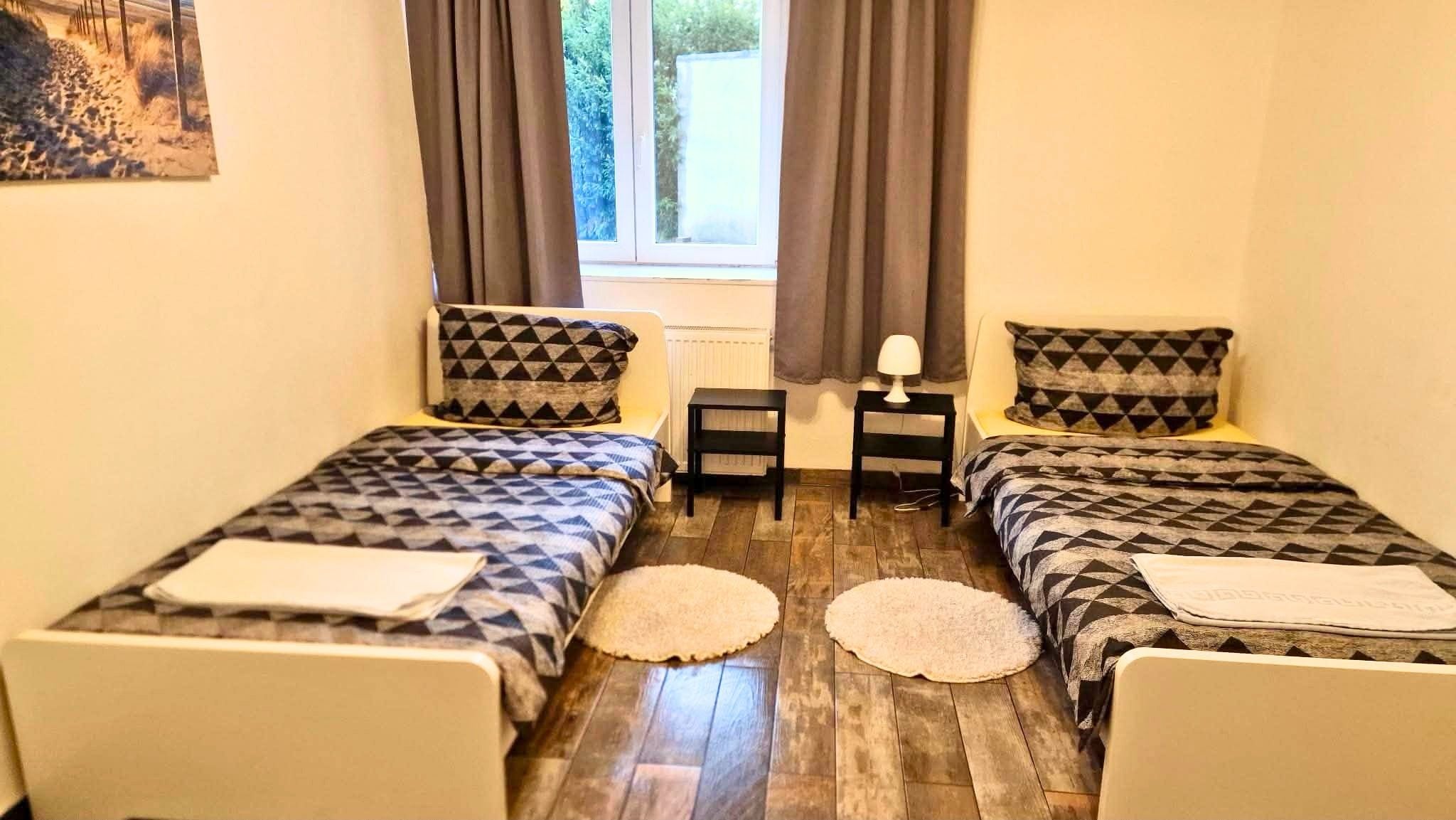 A room with two beds, a small table, a lamp, and a view of greenery outside.