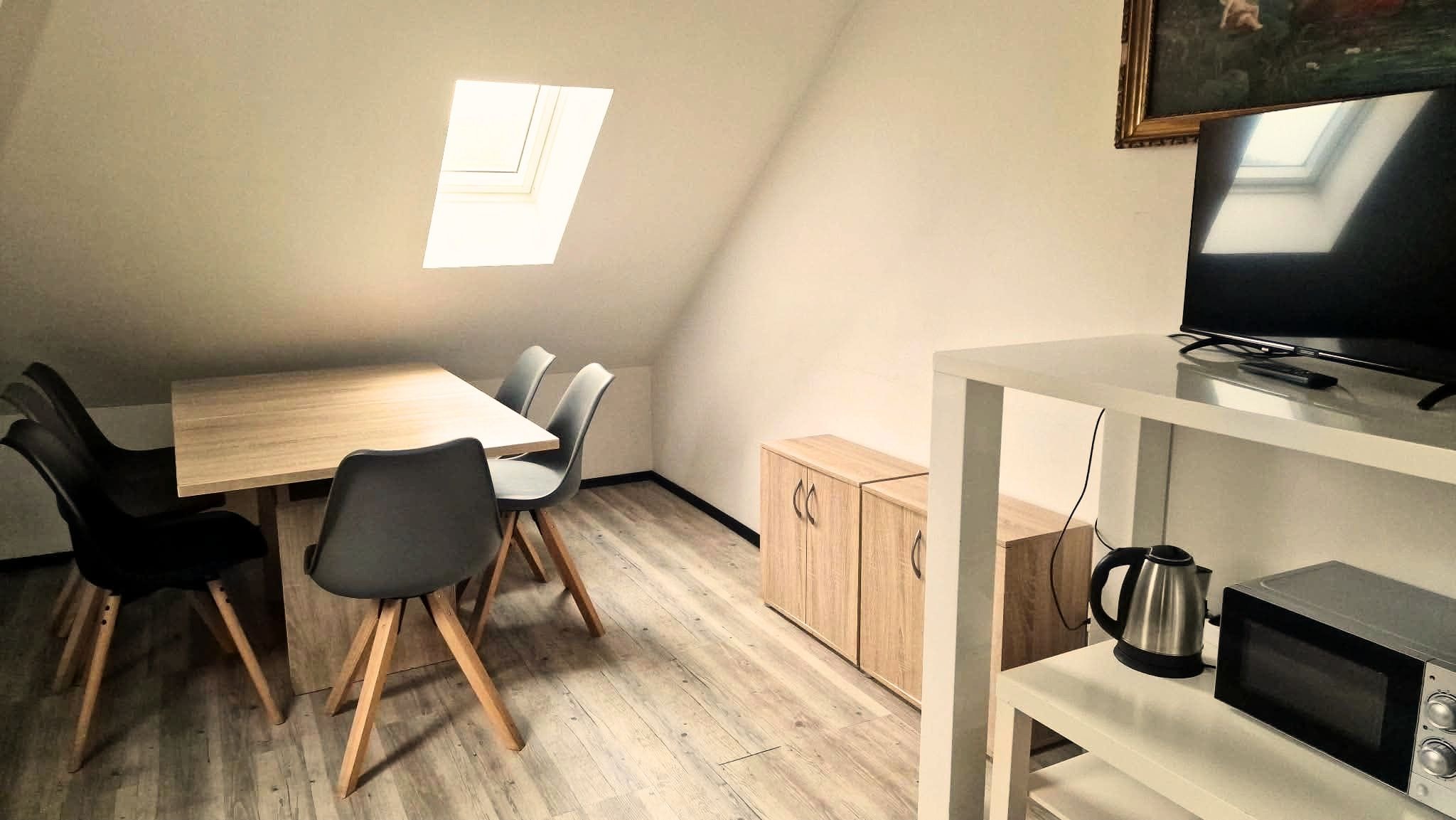 A room with a wooden dining table, grey chairs, a small cabinet, a television on a stand, and a kettle on a shelf.