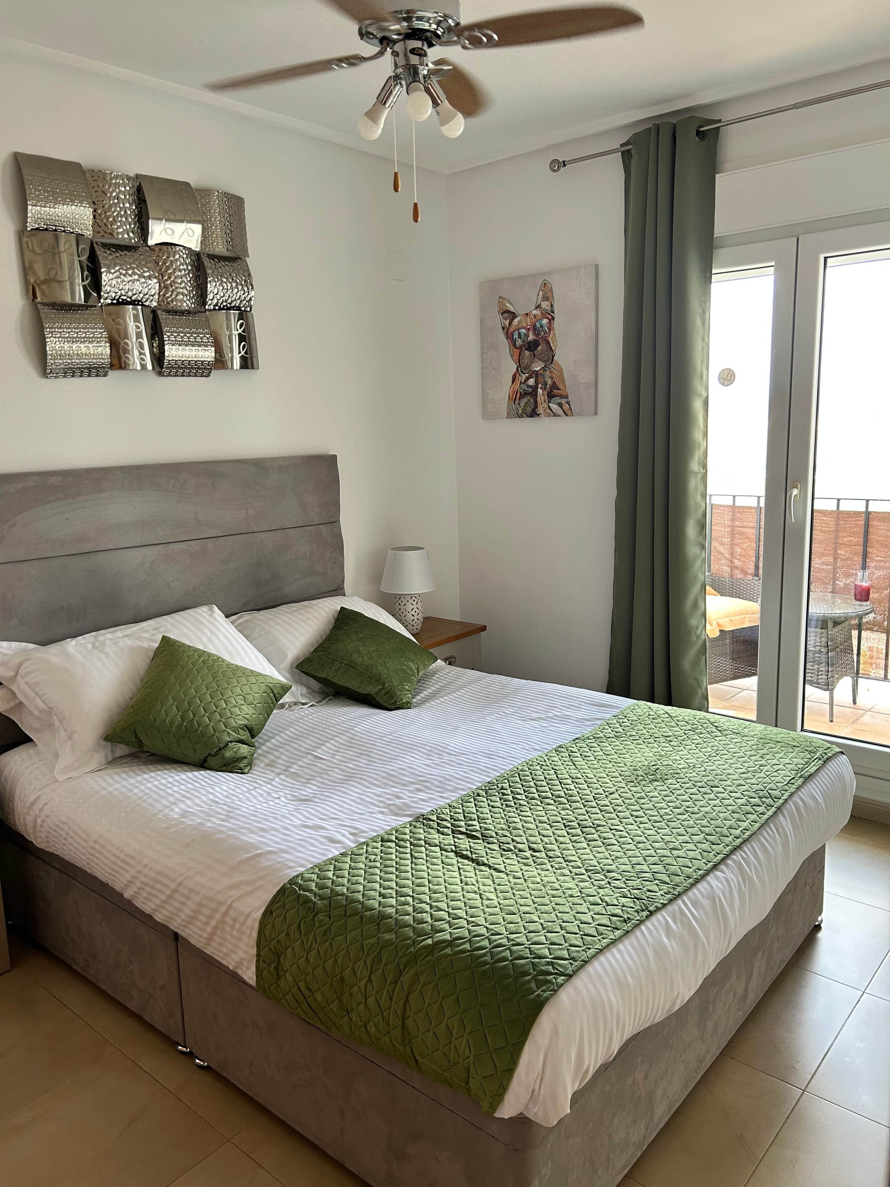 Une chambre à coucher avec un lit, un ventilateur de plafond, un tableau et une vue sur l’extérieur.