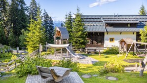 Una cabaña de madera rústica con techo inclinado, rodeada por un área verde con un parque infantil y una terraza de madera.