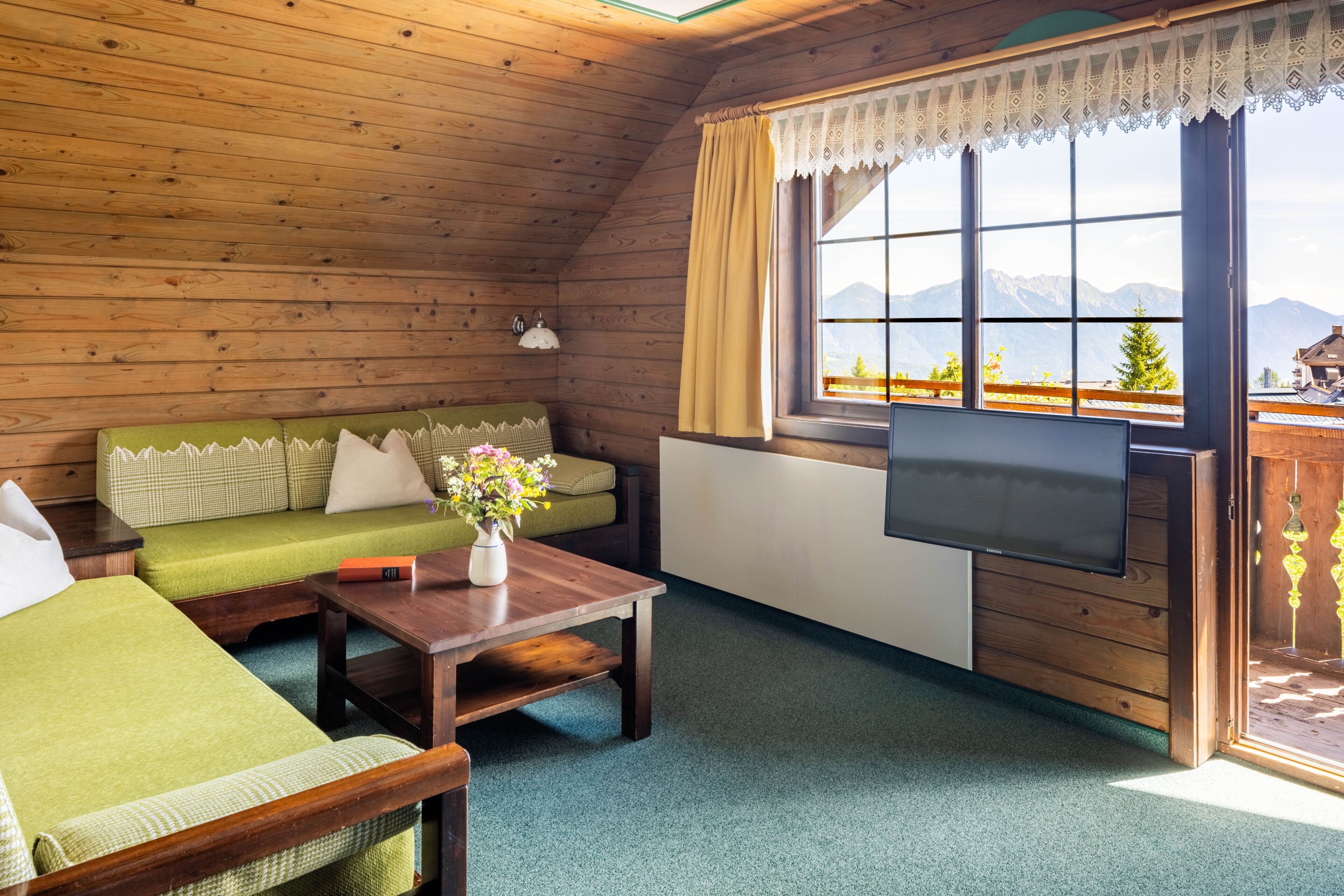 L’intérieur d’un chalet en bois, avec un canapé, une table ornée d’un vase, un téléviseur et une fenêtre donnant sur une vue sur les montagnes.