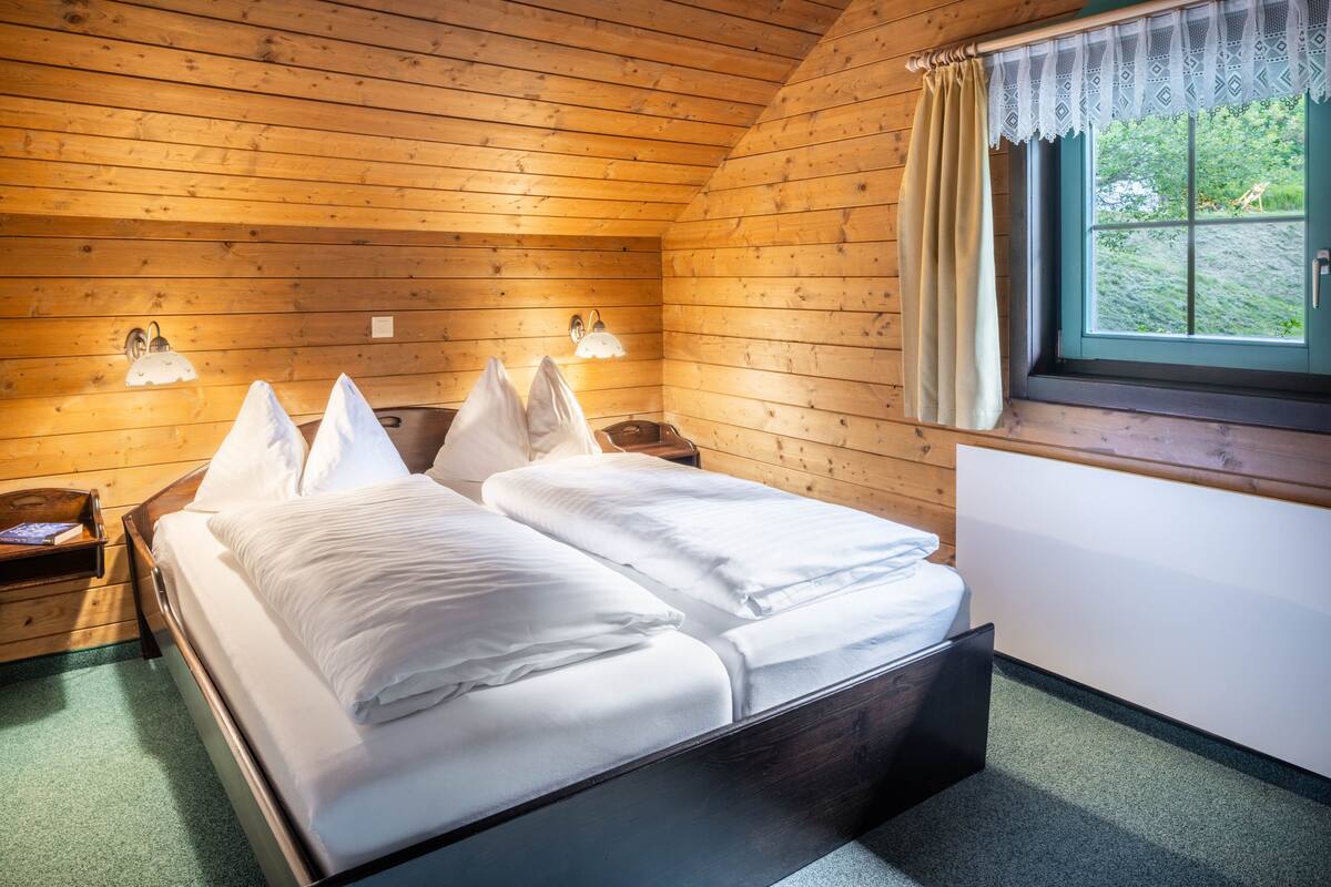 Habitación de cabaña de madera con una cama, dos lámparas fijadas a la pared, un ventanal con cortinas y vistas al verdor exterior.