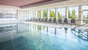 Una piscina interior con paredes de vidrio transparente, tumbonas y vistas a un lago de montaña.