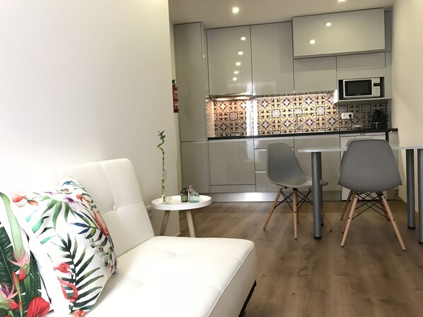 A compact living space with a white sofa, a small round table, and a kitchen area featuring a microwave and patterned backsplash. - Povoa do Mar (Povoa De Varzim)