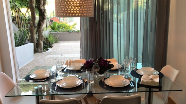 Mesa de jantar posta para seis com pratos, copos e talheres, com vista para um jardim.